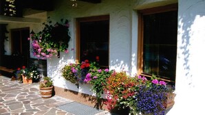 Un bâtiment aux murs blancs et aux nombreuses fenêtres. Des jardinières contenant différentes sortes de fleurs ornent l’extérieur.