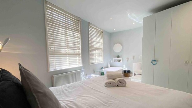 A bedroom with a bed, two windows with blinds, a bedside table with towels, a round mirror, and a wardrobe. - Charming 2-bed Apt With 2 Bathtub Central London (London)