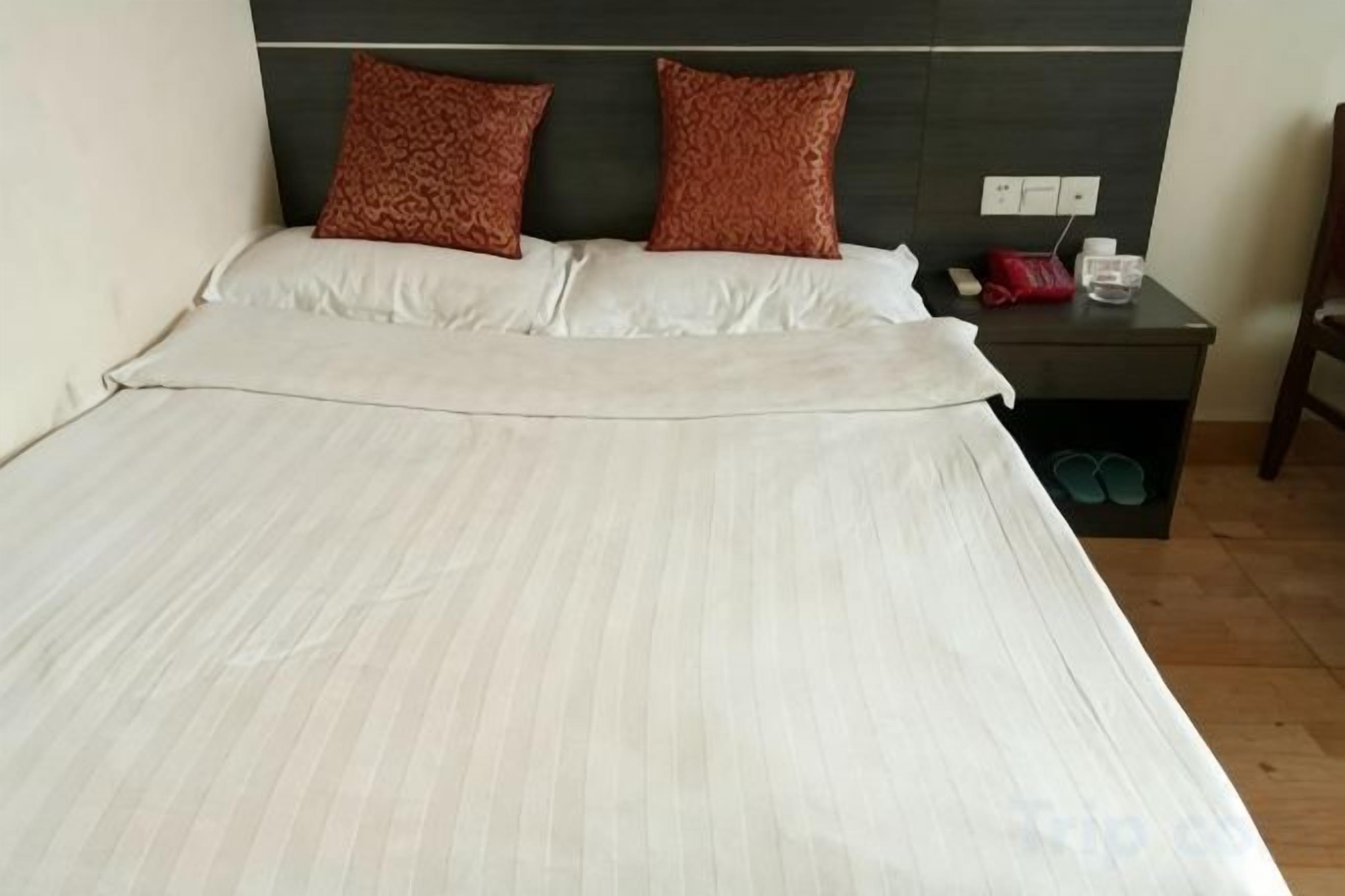 Una cama con una colcha blanca y dos almohadas marrones, una mesita de noche con una botella roja y un recipiente blanco, y un par de zapatos debajo de la mesita de noche.