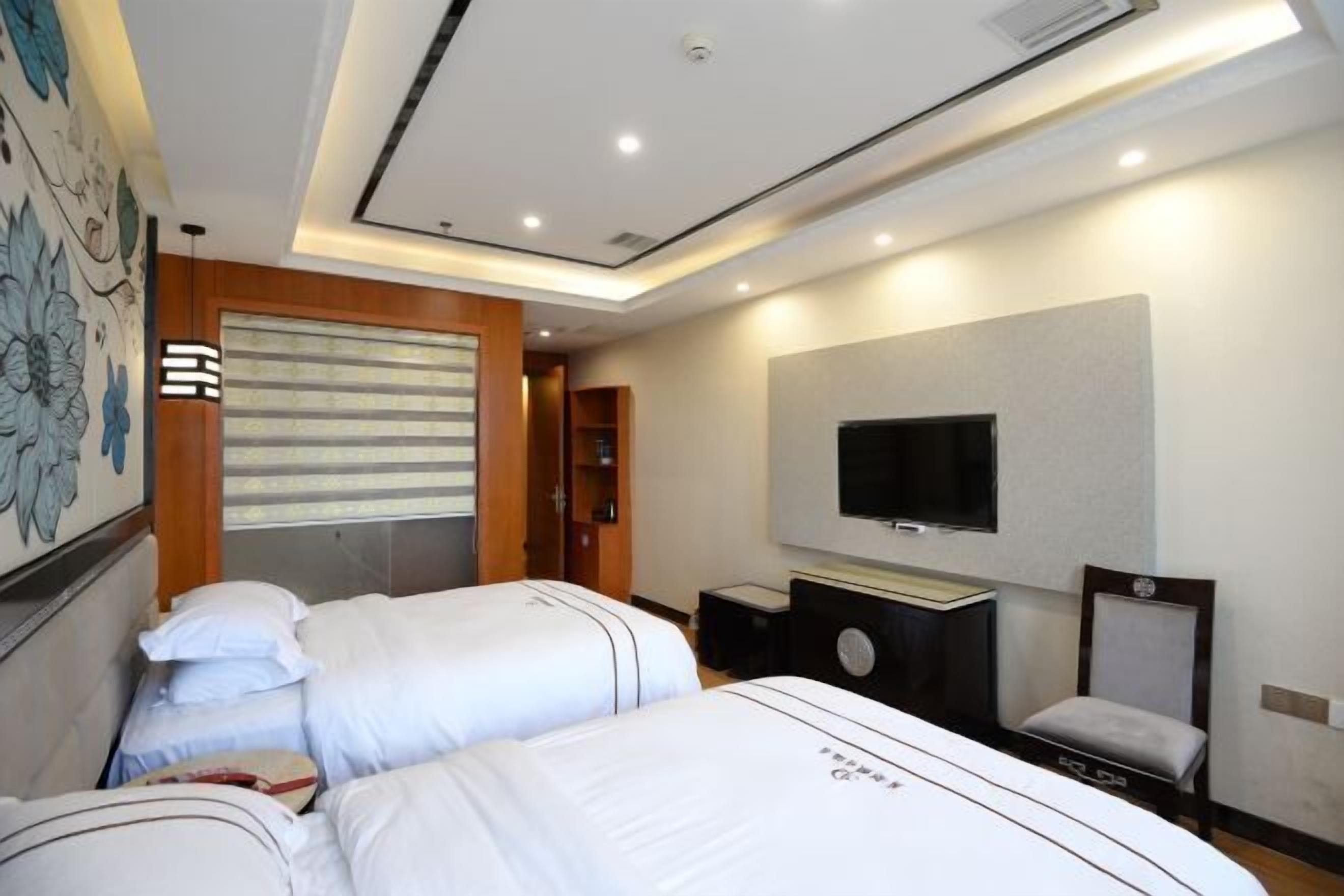A hotel room with two beds, a flat-screen TV mounted on the wall, a desk with a chair, and a large window with blinds.