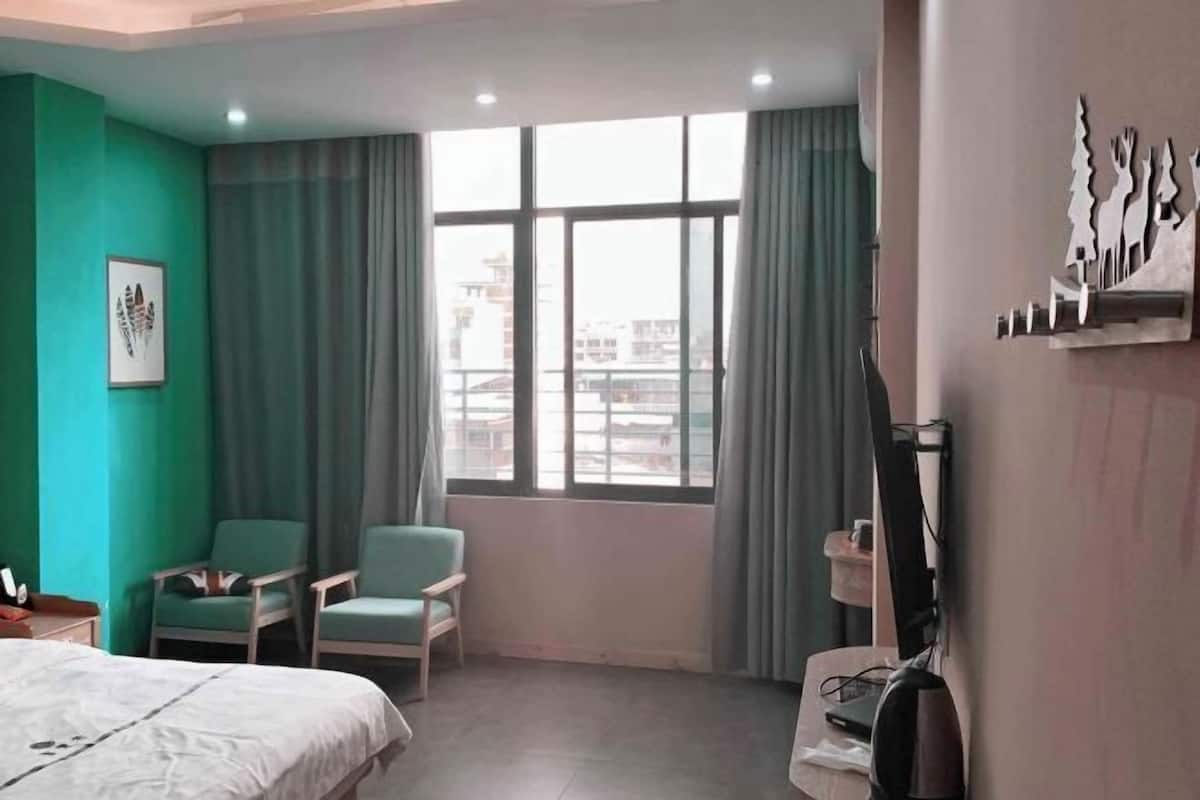 A hotel room with a bed, two chairs, a window with curtains, a wall-mounted shelf, and a view of buildings outside.