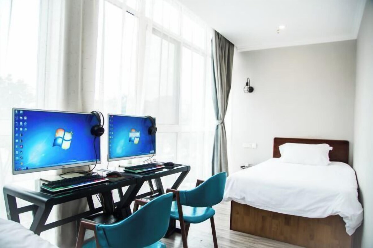 A hotel room with two monitors on a desk, a bed, and a window with curtains.