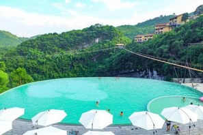 A large outdoor swimming pool surrounded by white umbrellas, set against a backdrop of lush green hills and a rope bridge. - Cotton Tiankeng Hotel (Luocheng)