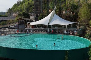 A large outdoor swimming pool with a white canopy, surrounded by a rocky landscape and a small building. - Cotton Tiankeng Hotel (Luocheng)
