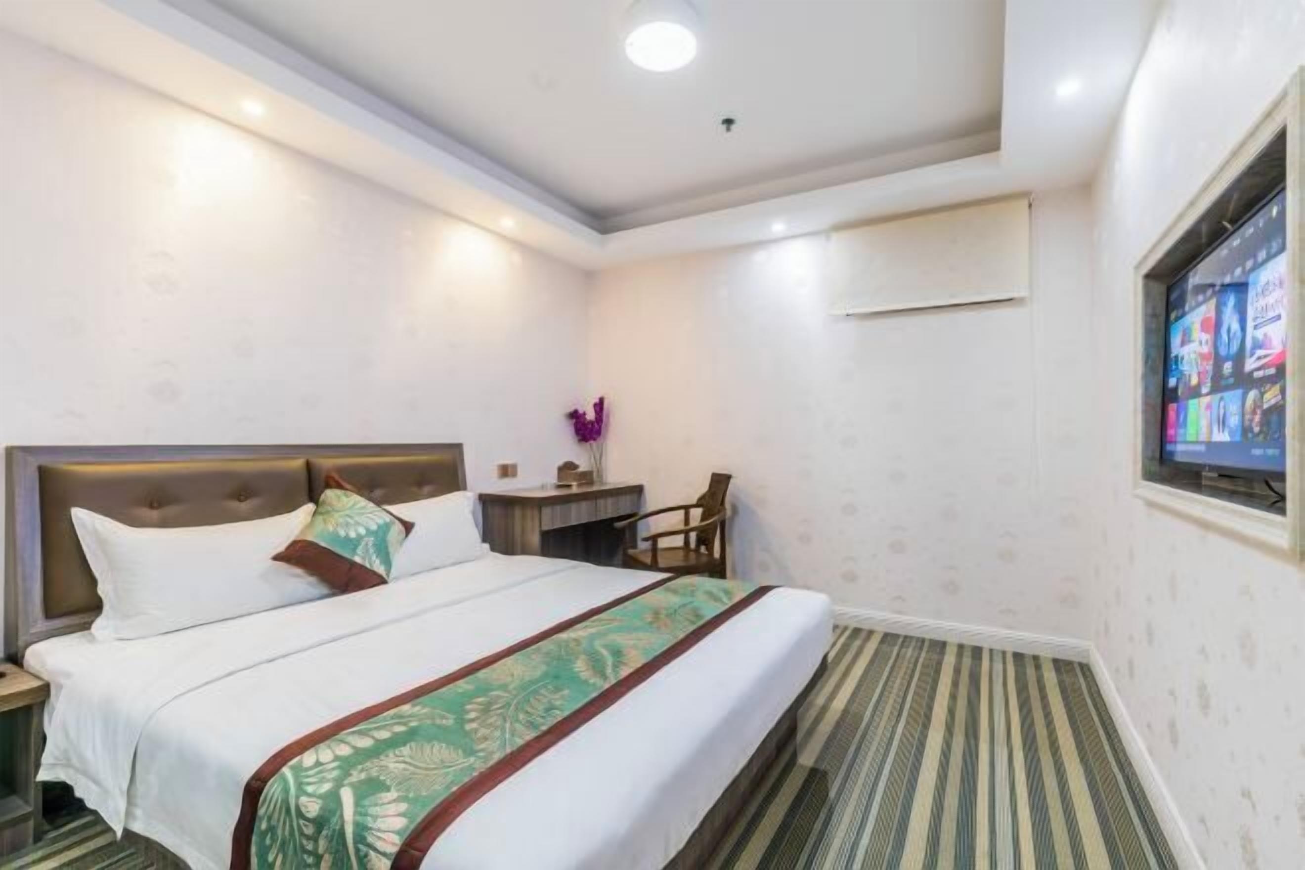 A hotel room with a bed, a desk, a chair, a TV, and an air conditioning unit.