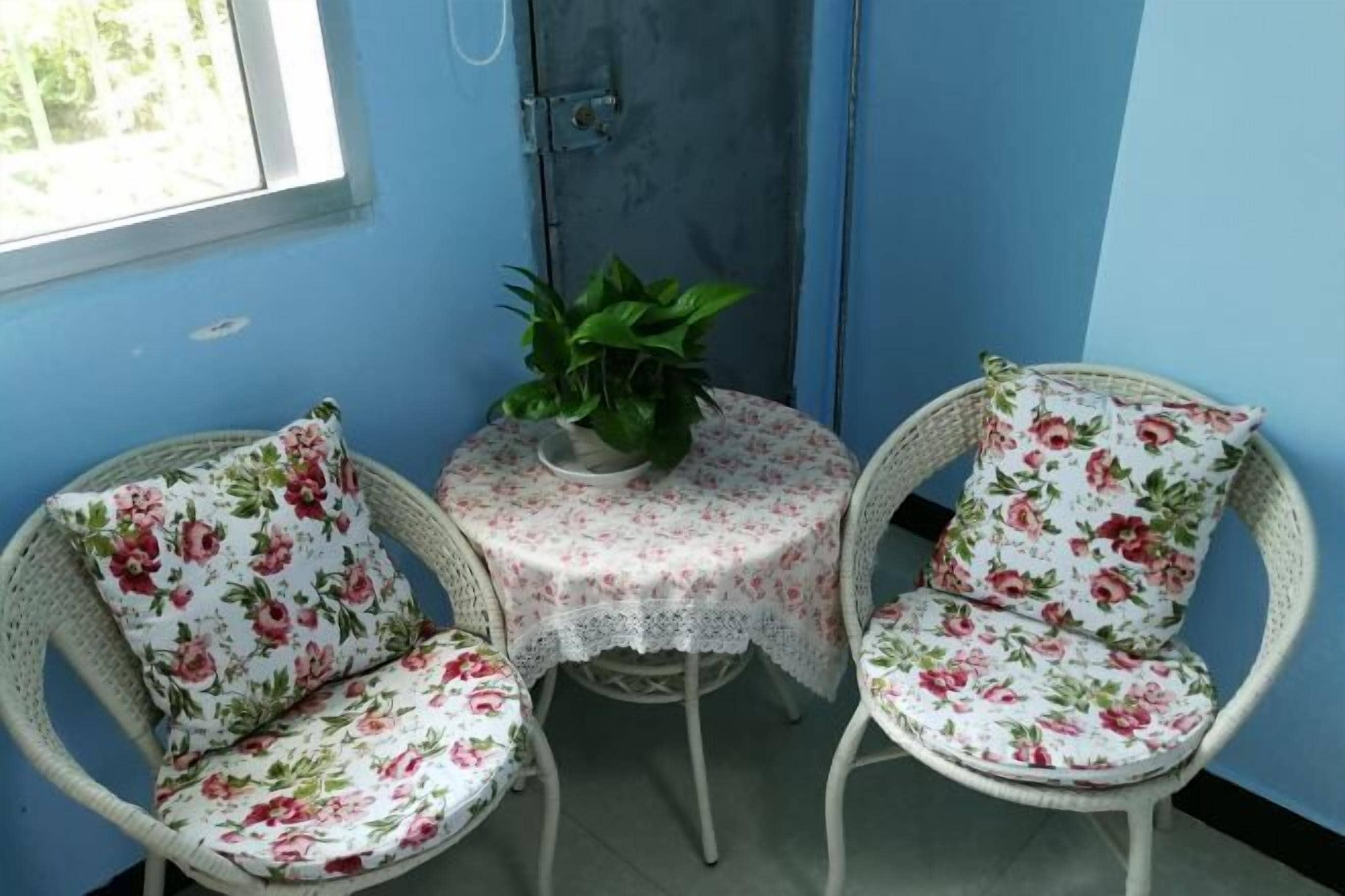 Una habitación de esquina con dos sillones de mimbre, una pequeña mesa redonda con mantel de flores y una planta en maceta.