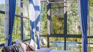 Un lit avec des oreillers et une couverture, de grandes fenêtres avec des rideaux et une vue sur la verdure à l’extérieur.