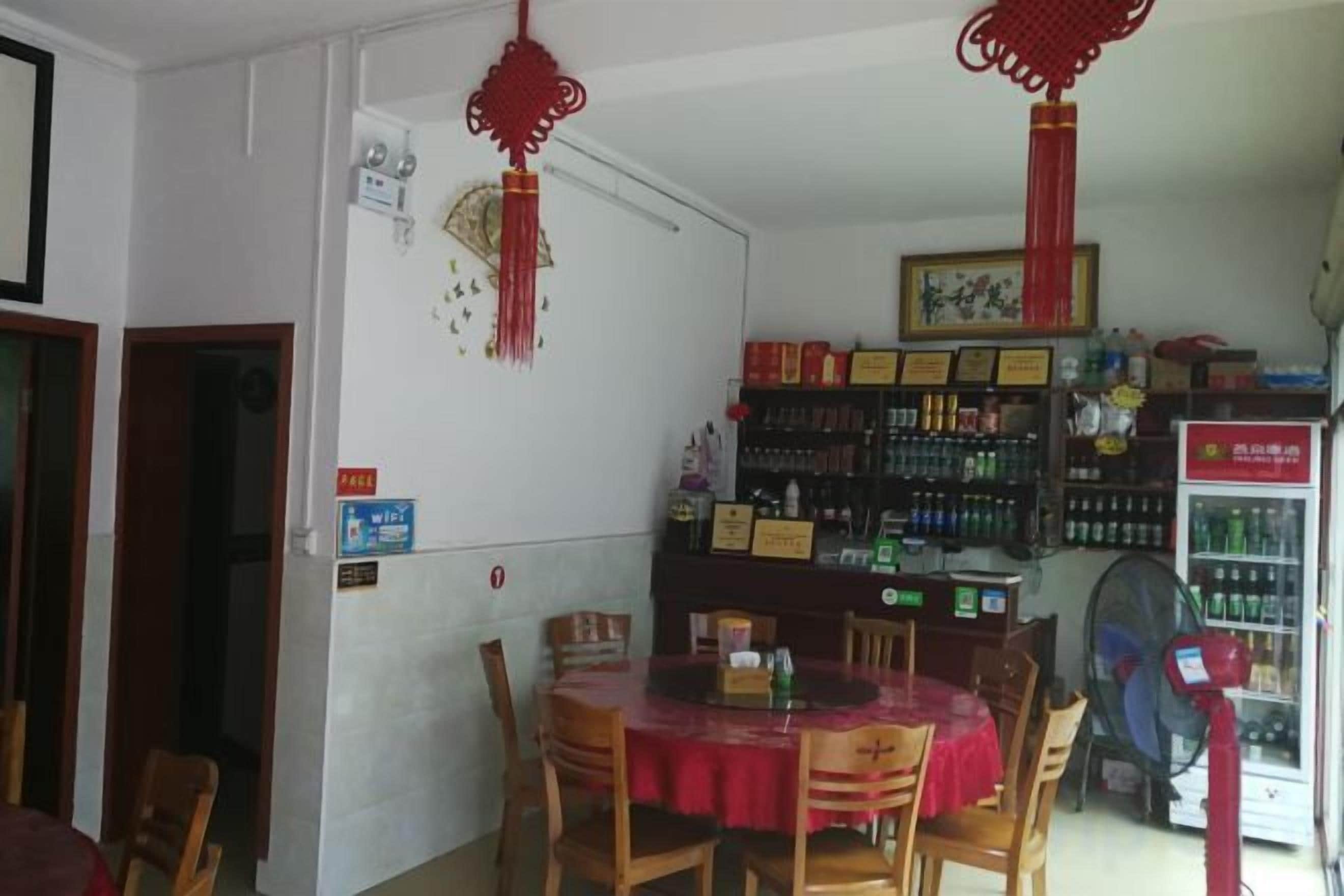 Un área de comedor con una mesa redonda, sillas y un mantel rojo. Se ven estantes con diversos objetos y un refrigerador al fondo.