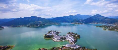 Vista aérea de un complejo turístico a orillas de un lago, con una isla central que presenta varios edificios, rodeado de montañas y aguas cristalinas.