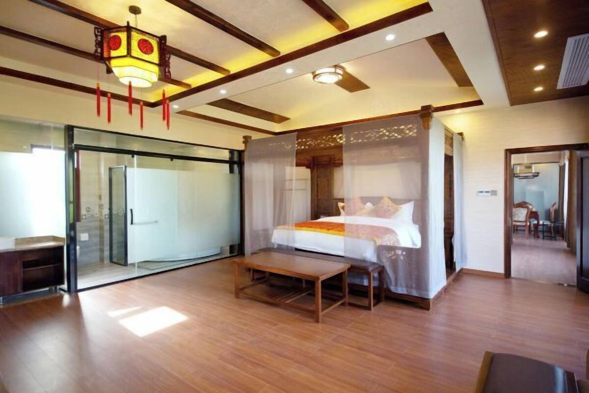 Een slaapkamer met een bed, nachtkastjes, een bankje, een glazen deur, en een plafond met houten balken en inbouwverlichting.