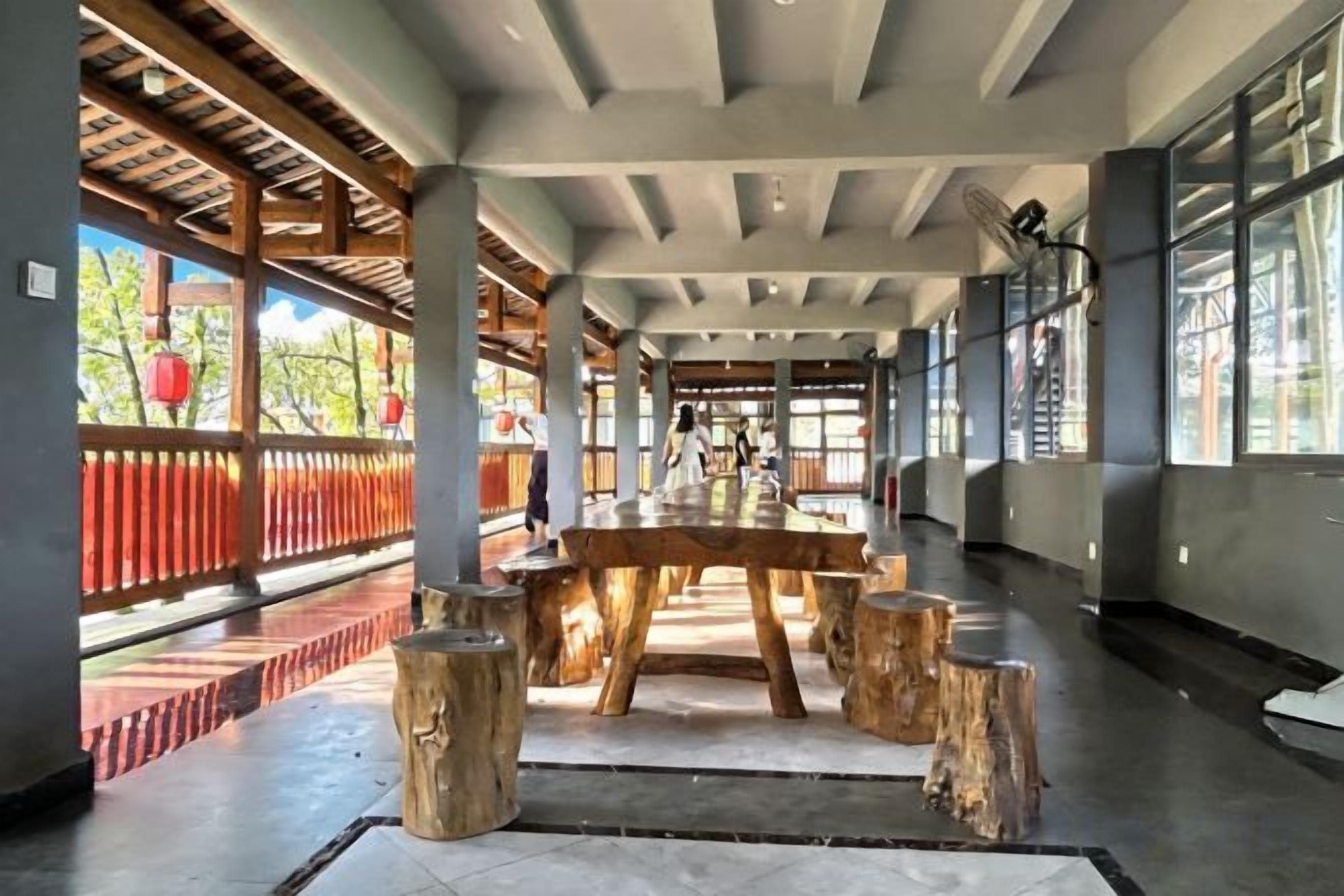 Un interior espacioso con una gran mesa de madera rodeada de taburetes, un techo alto con vigas a la vista y ventanas amplias que dejan entrar la luz natural.