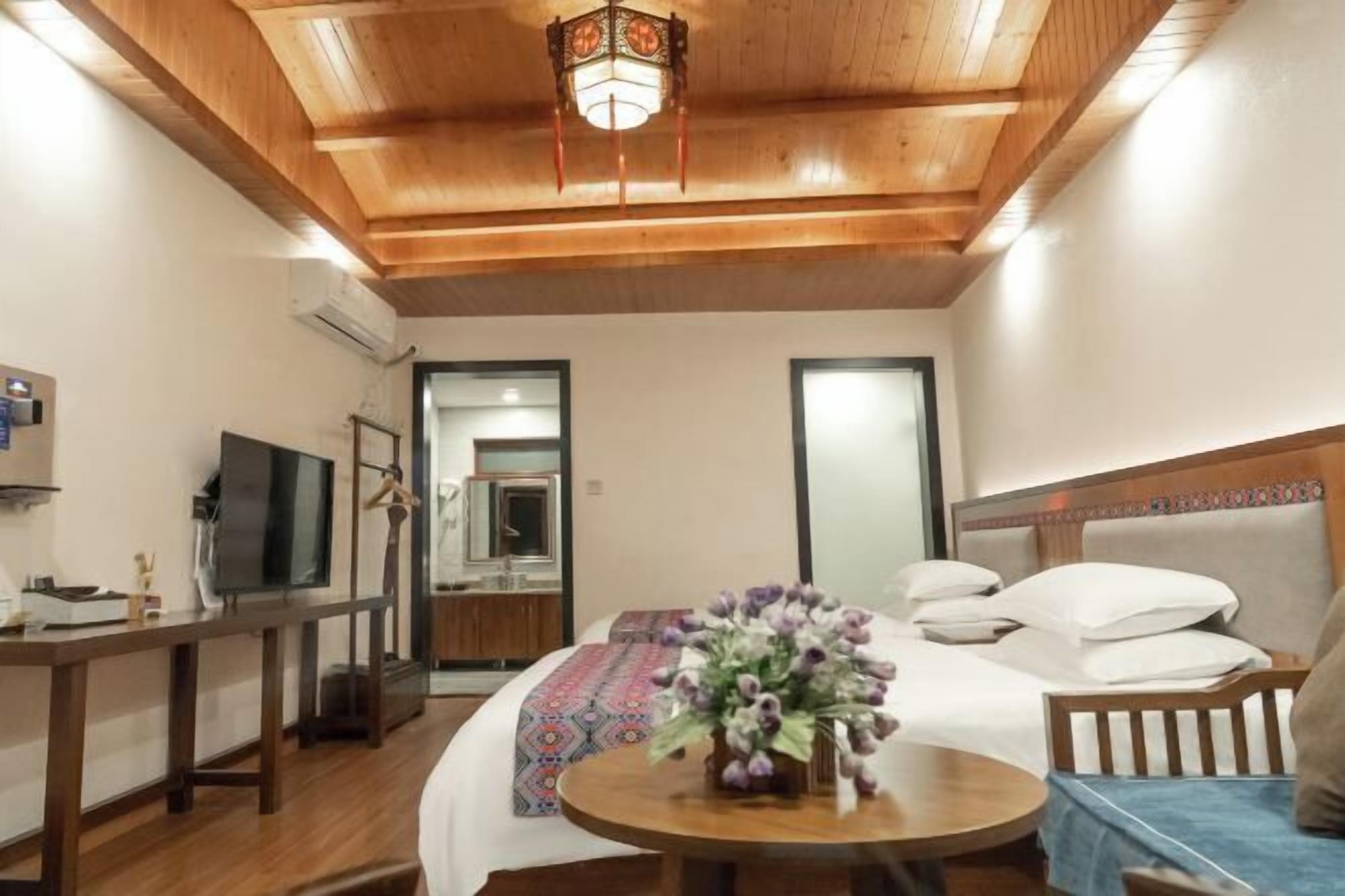 Habitación de hotel con techo de madera, cama con edredón estampado, mesa redonda con un jarrón de flores y televisor sobre un mueble.