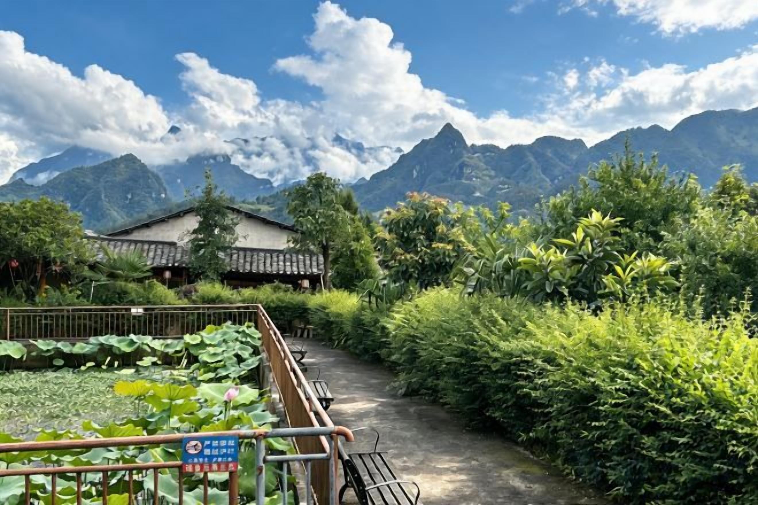 Un camino que lleva a una edificación tradicional rodeada de abundante vegetación y montañas al fondo.