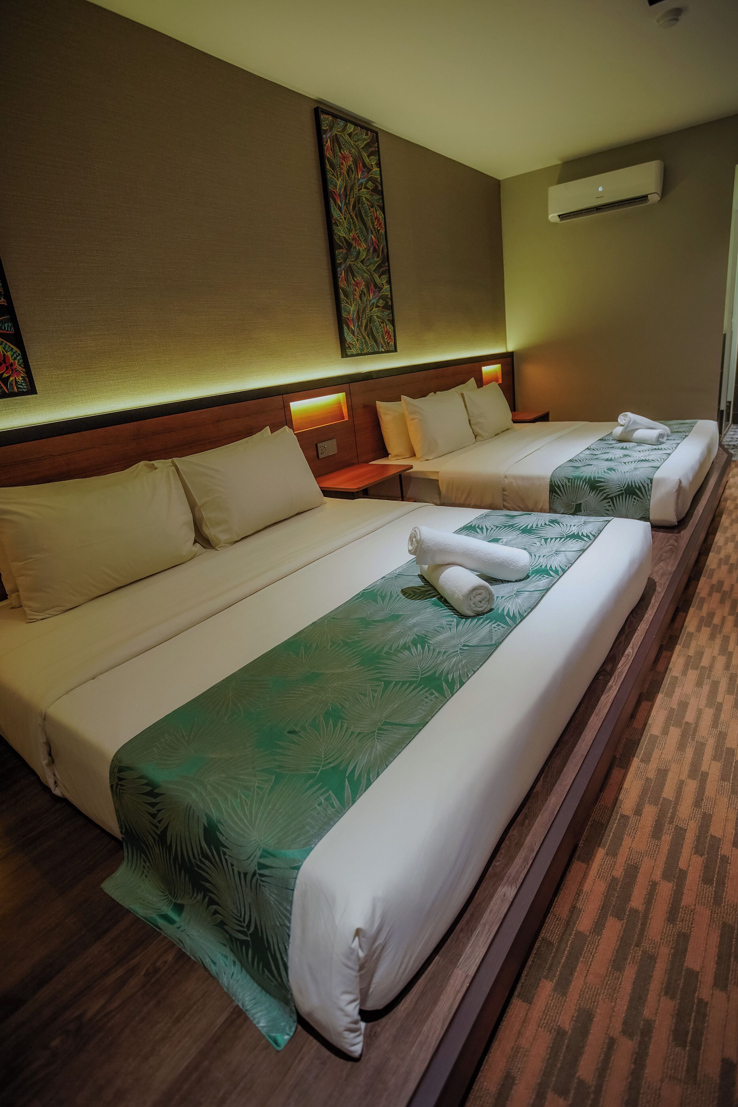 Habitación de hotel con dos camas, cada una con cubrecama estampado con hojas verdes, almohadas blancas y toallas dobladas con cuidado.