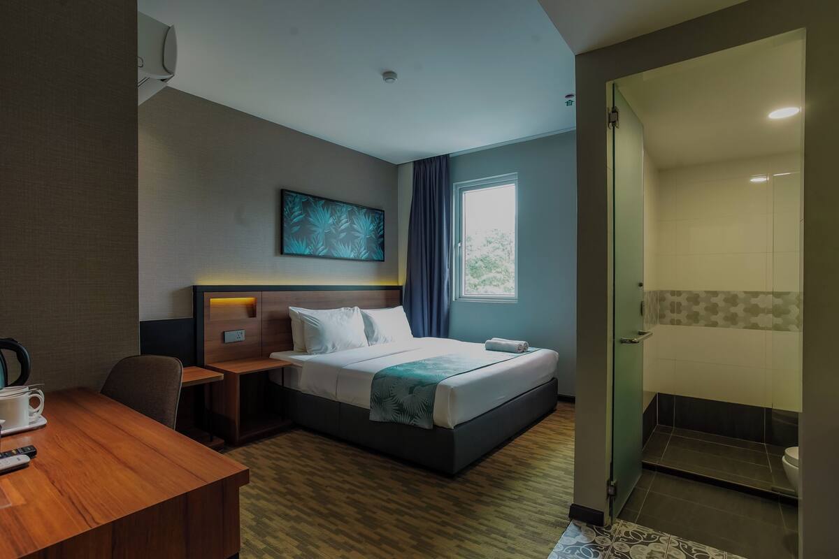 A hotel room with a bed, a desk, a chair, a window with curtains, a shower area, and a wall-mounted artwork.