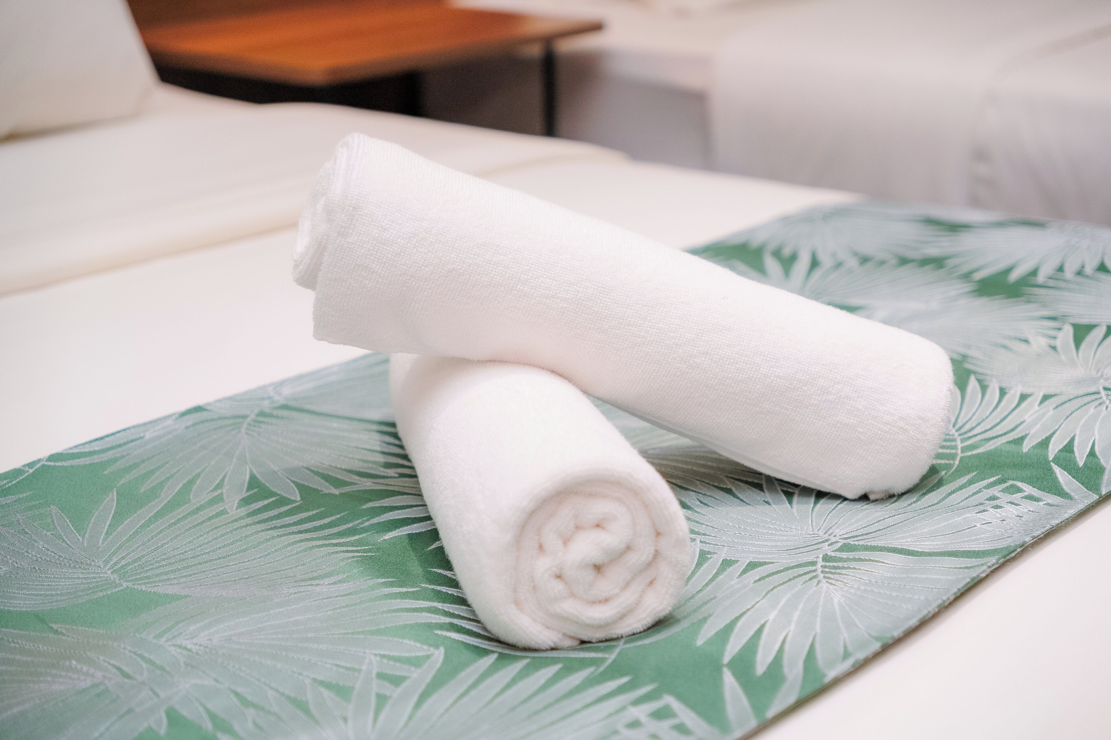 Una toalla blanca doblada con cuidado sobre un tapete con estampado de hojas verdes, colocada en una cama de habitación de hotel.