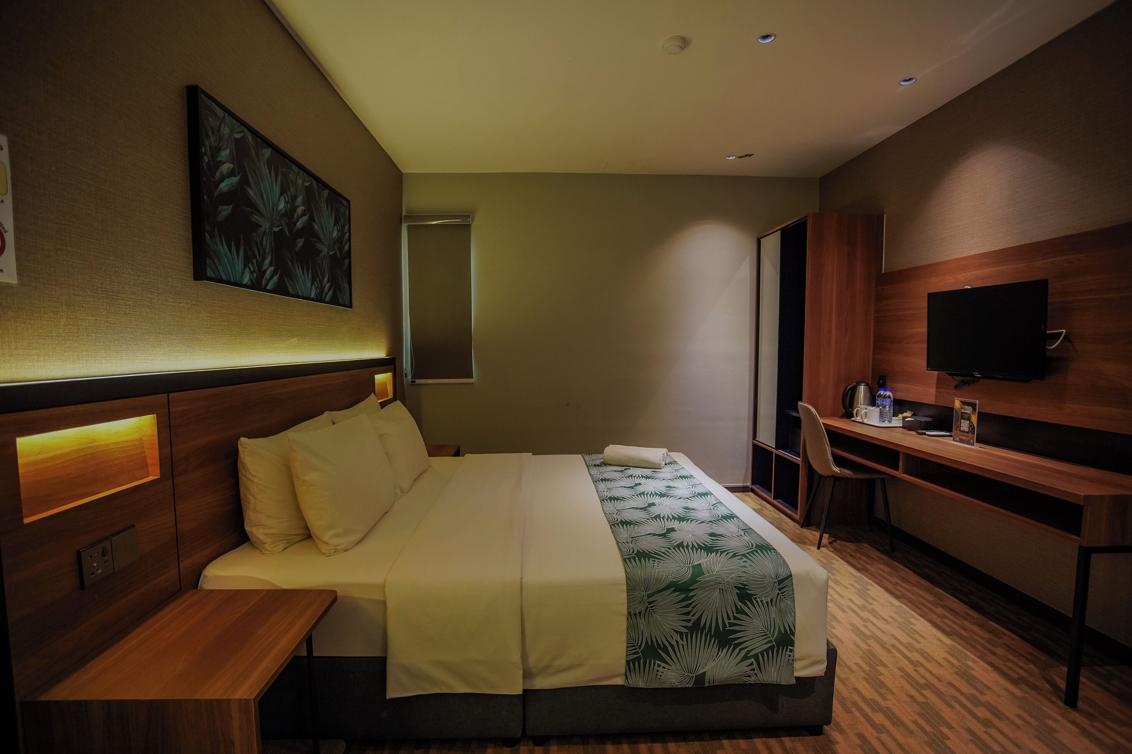 A hotel room with a bed, bedside table, desk, chair, TV, and a painting on the wall.