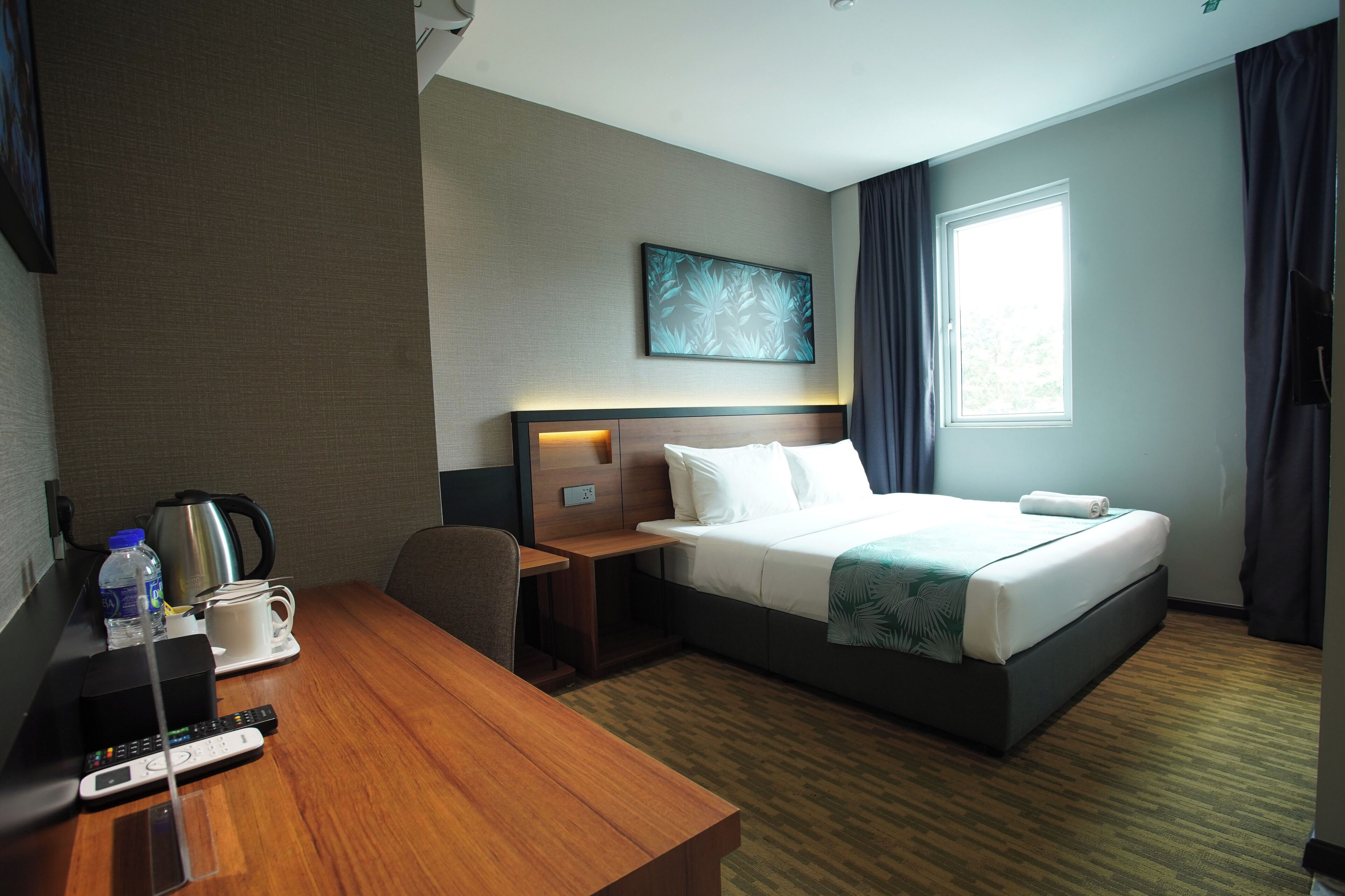 A hotel room with a bed, a desk, a chair, a coffee maker, a remote control, and a window with curtains.