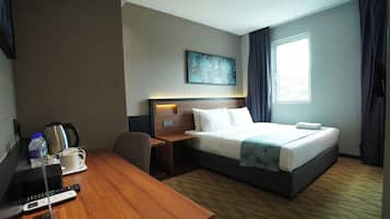 A hotel room with a bed, a desk, a chair, a coffee maker, a remote control, and a window with curtains.