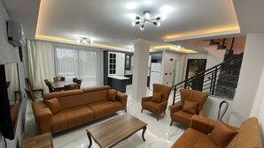 Uma sala de estar moderna com sofá seccional, poltronas e uma mesa de centro. Há uma área de jantar com mesa e cadeiras, e uma cozinha ao fundo.