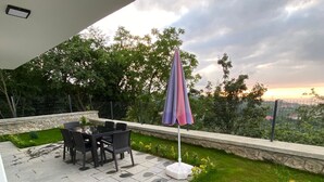 Um pátio ao ar livre com mesa e cadeiras de pedra, gramado com flores amarelas e vista para árvores e um céu nublado.