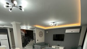 Uma sala de estar moderna com área de jantar, sofás cinzentos, uma mesa branca e uma TV fixada na parede.