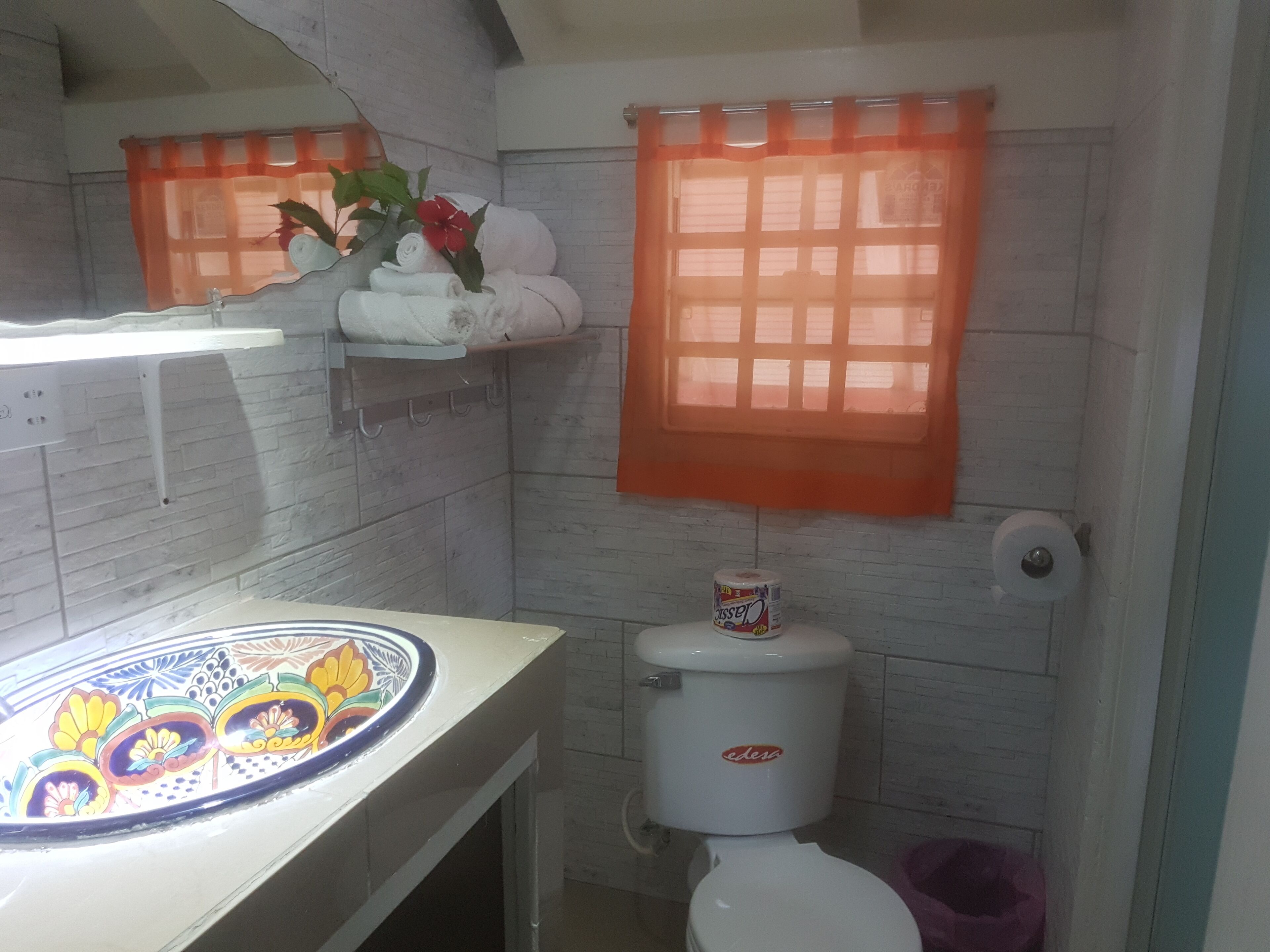 A bathroom with a toilet, a sink with a decorative plate, a towel rack with towels, and a window with an orange curtain.