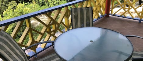 Un balcon avec une table en verre et des chaises à rayures, donnant sur un espace verdoyant luxuriant.