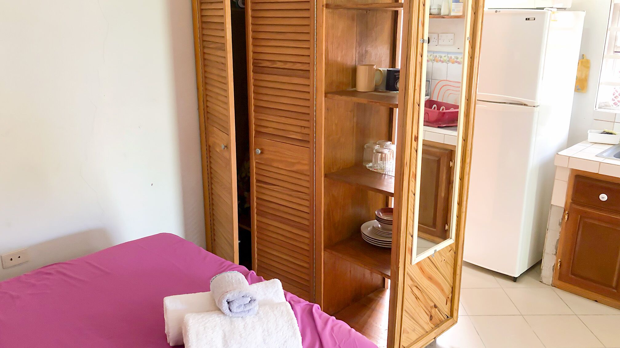 A room with a bed, a wooden wardrobe with a mirror, a kitchen area with a refrigerator, and a sink.