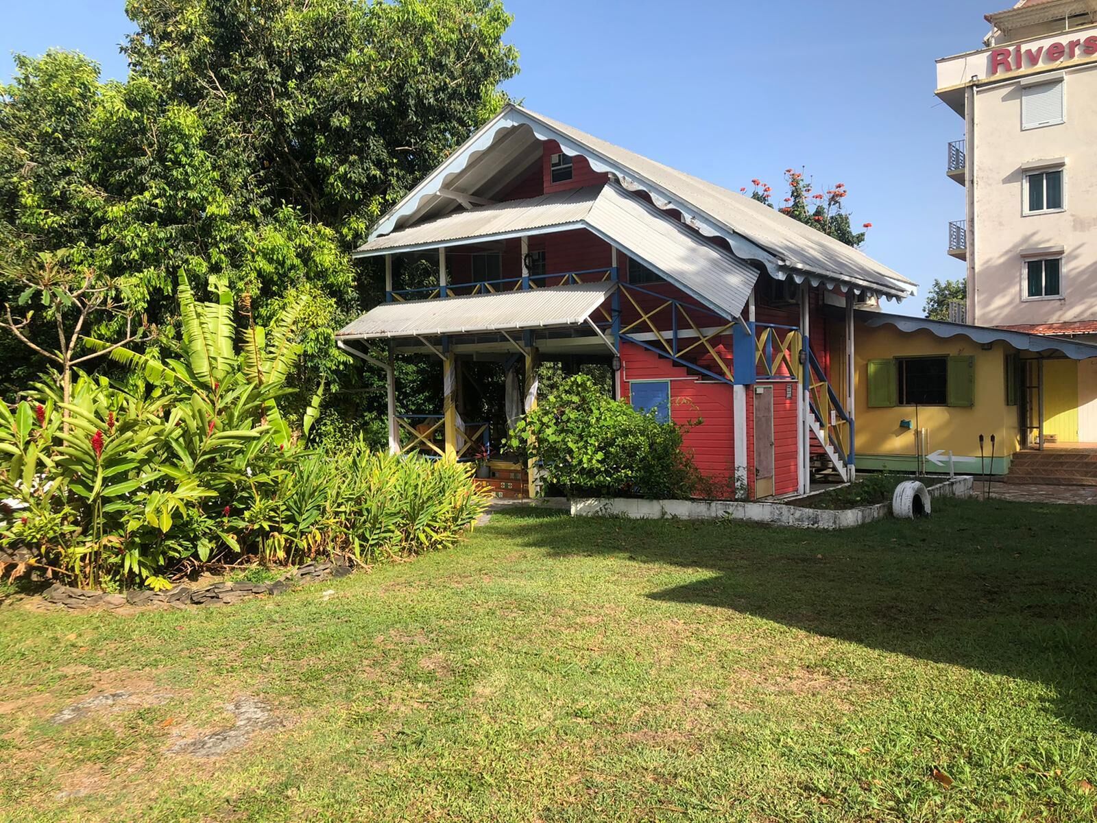 A two-story building with a red and white exterior, a covered porch, and a small garden area with tropical plants.