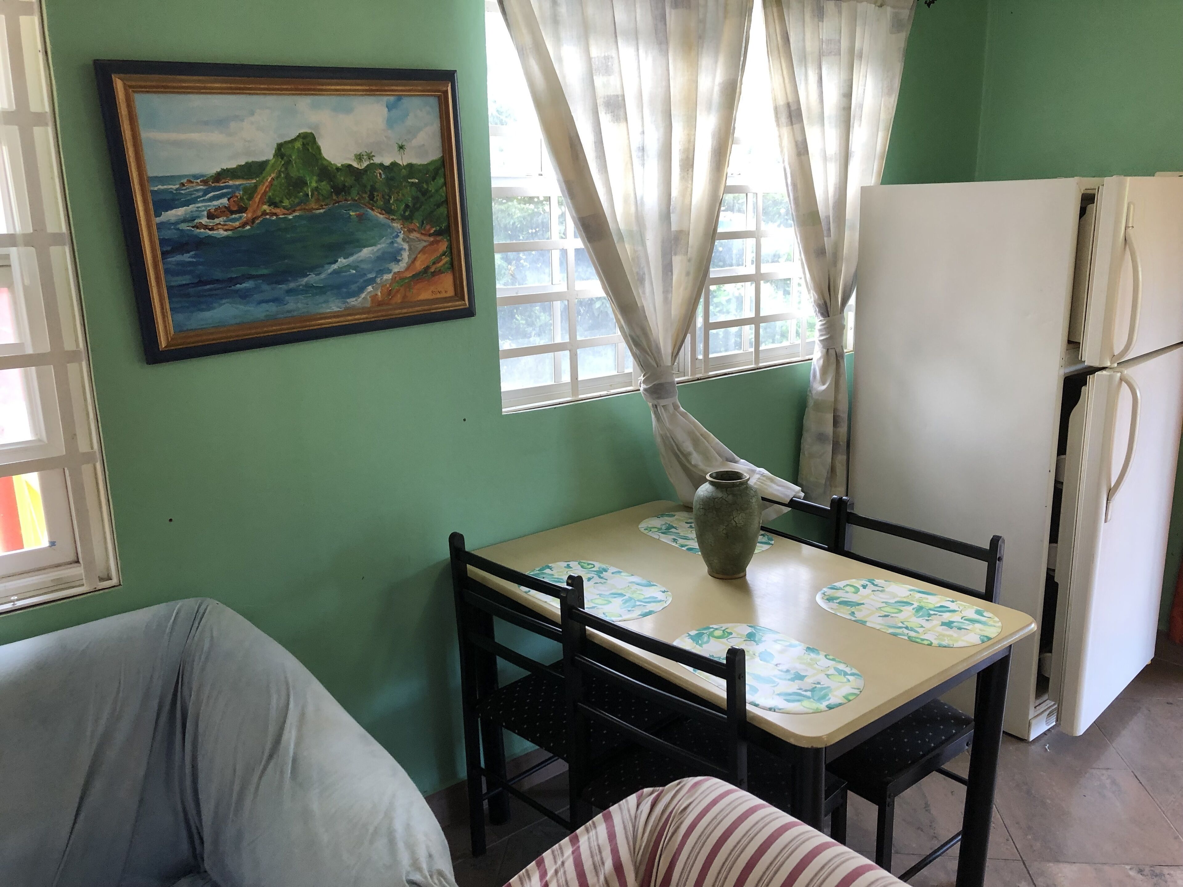 A room with a table, chairs, a refrigerator, a painting, and a window with curtains.