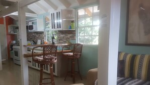 A kitchen with white cabinets, a dining area with wicker chairs, and a view of the outdoors through a window. - Coco Mango Suites (Portsmouth)
