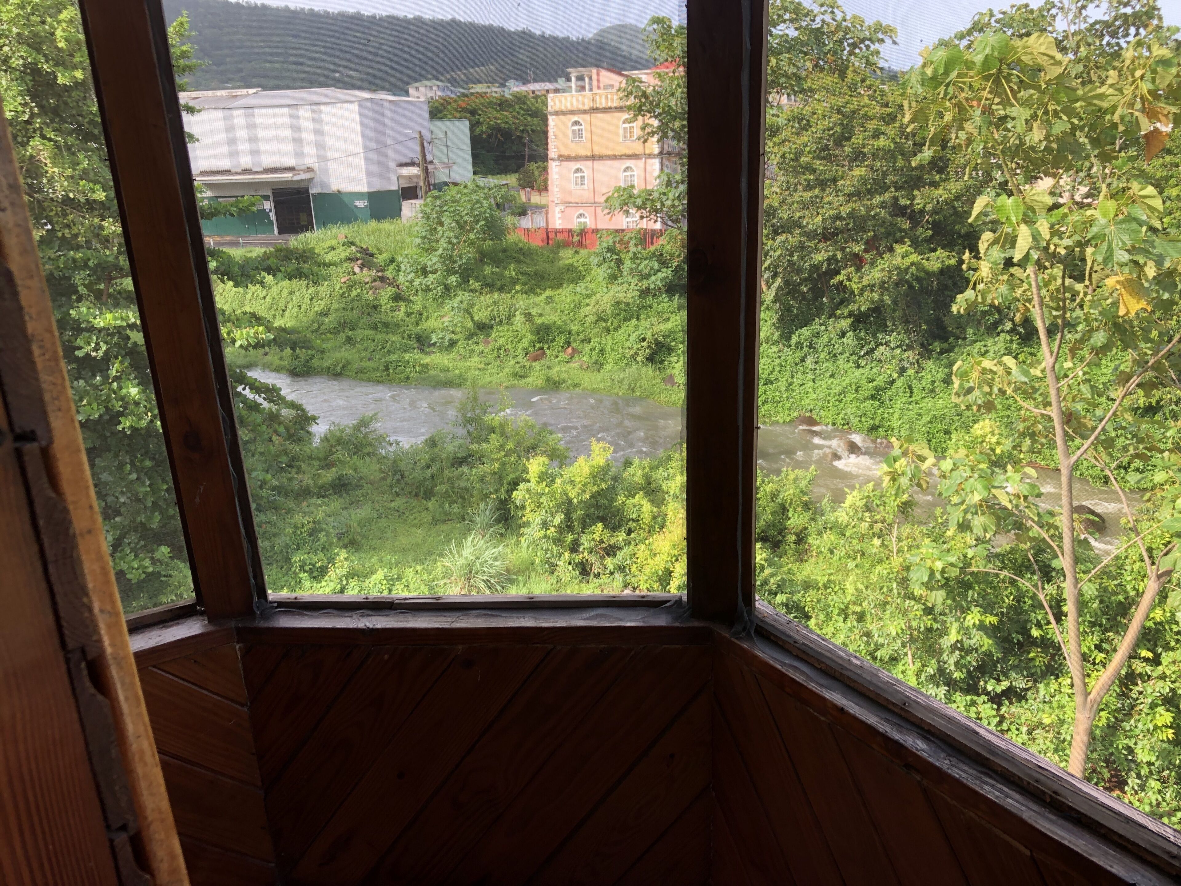 Vista pela janela de um quarto com decoração em madeira, com vista para um rio, vegetação e edifícios ao fundo.
