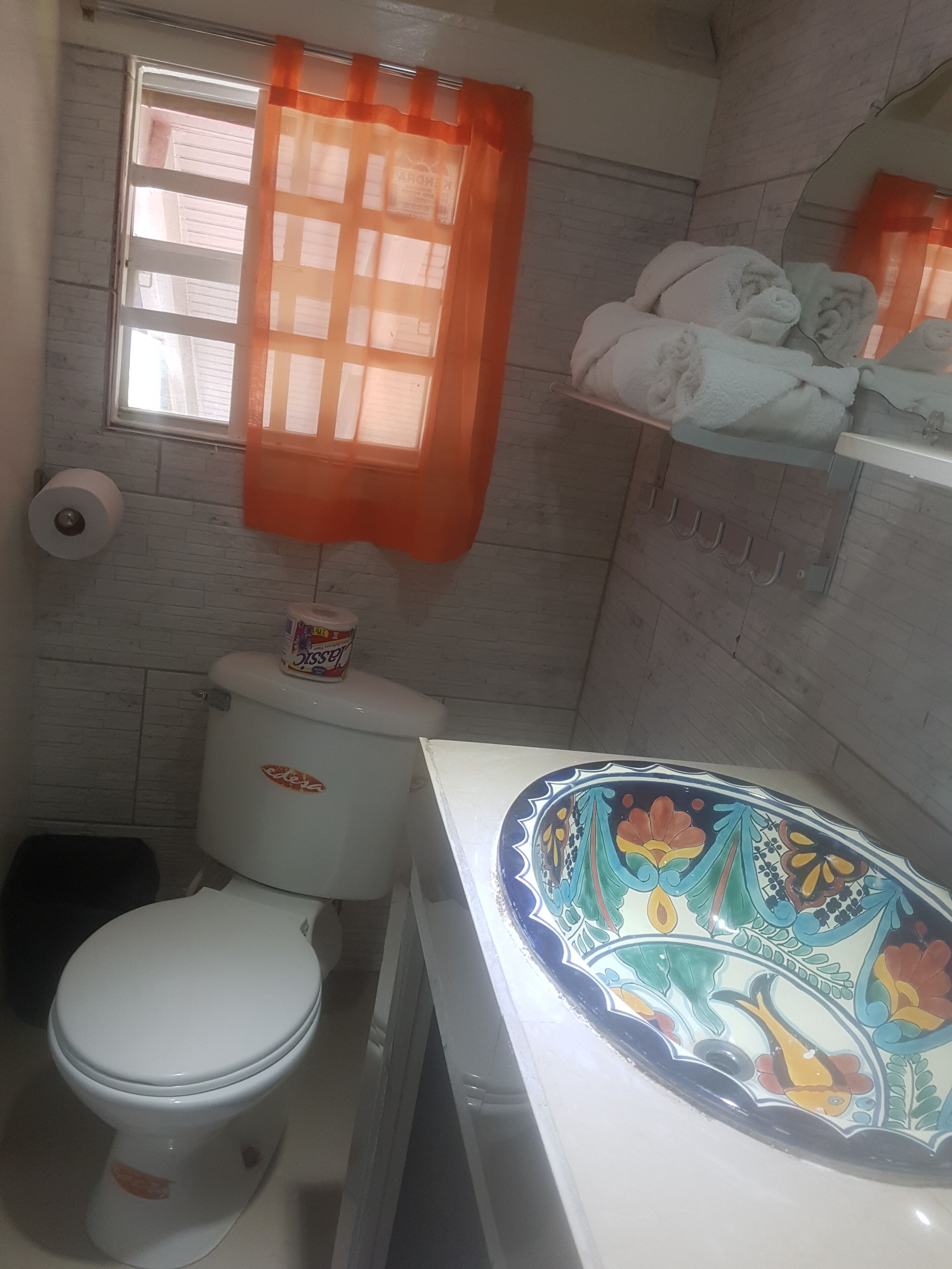 A small bathroom with a toilet, a roll of toilet paper, a shelf with towels, and a window with an orange curtain.