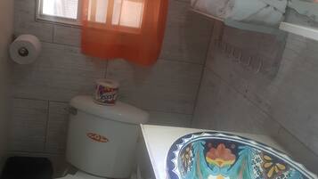 A small bathroom with a toilet, a roll of toilet paper, a shelf with towels, and a window with an orange curtain.