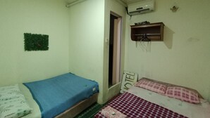 A small room with two beds, one with a blue mattress and the other with a red and white checkered mattress. There is a small shelf with items on it, a wall-mounted air conditioner, and a door leading to another room.