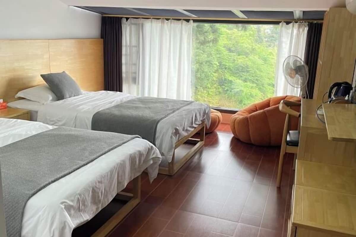 Quarto de hotel com duas camas, cabeceira de madeira, escrivaninha com cadeira, janela com cortinas e um ventilador.