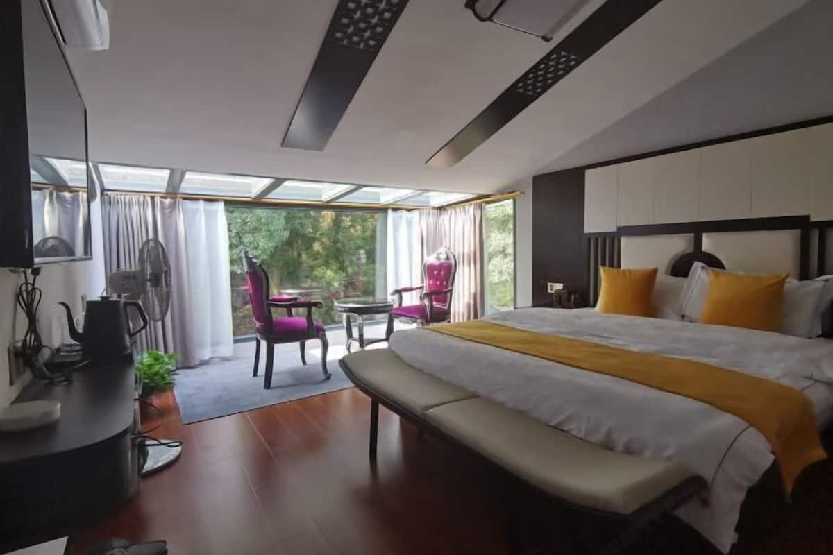 Quarto de hotel moderno com uma cama grande, um banco, uma mesa com chaleira e vista para o verde através de janelas amplas.