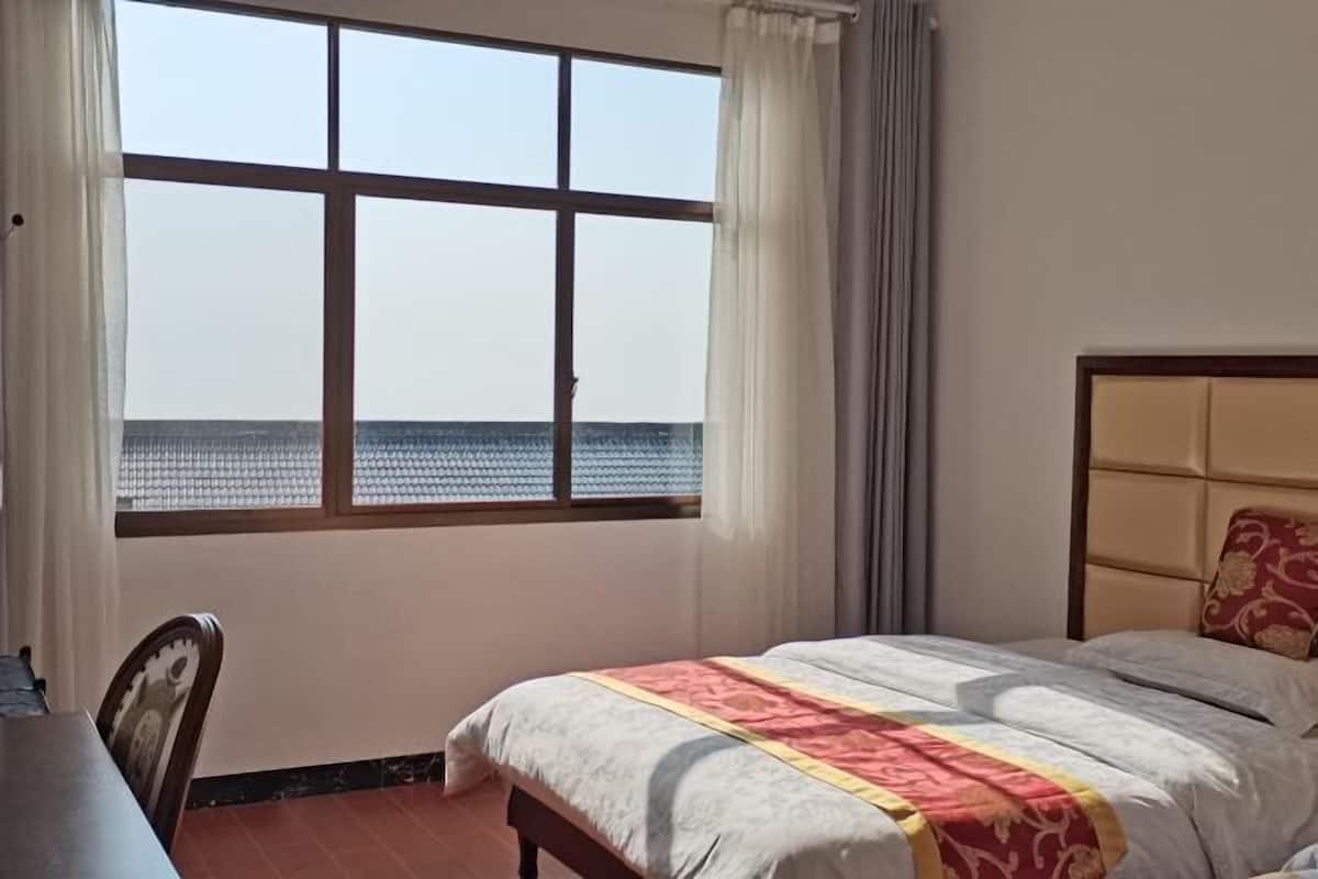Quarto de hotel com uma janela ampla que oferece vista para o mar, uma cama com colcha estampada e uma mesa de cabeceira de madeira.