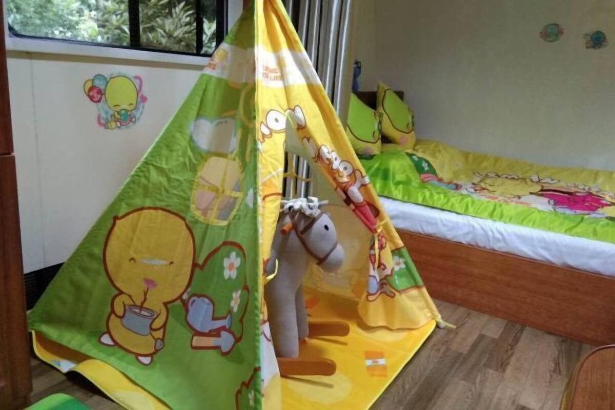 Een kinderspeeltent met tekenfilmfiguren, een bed met een groen-gele dekbedovertrek, en een raam met uitzicht op bomen.