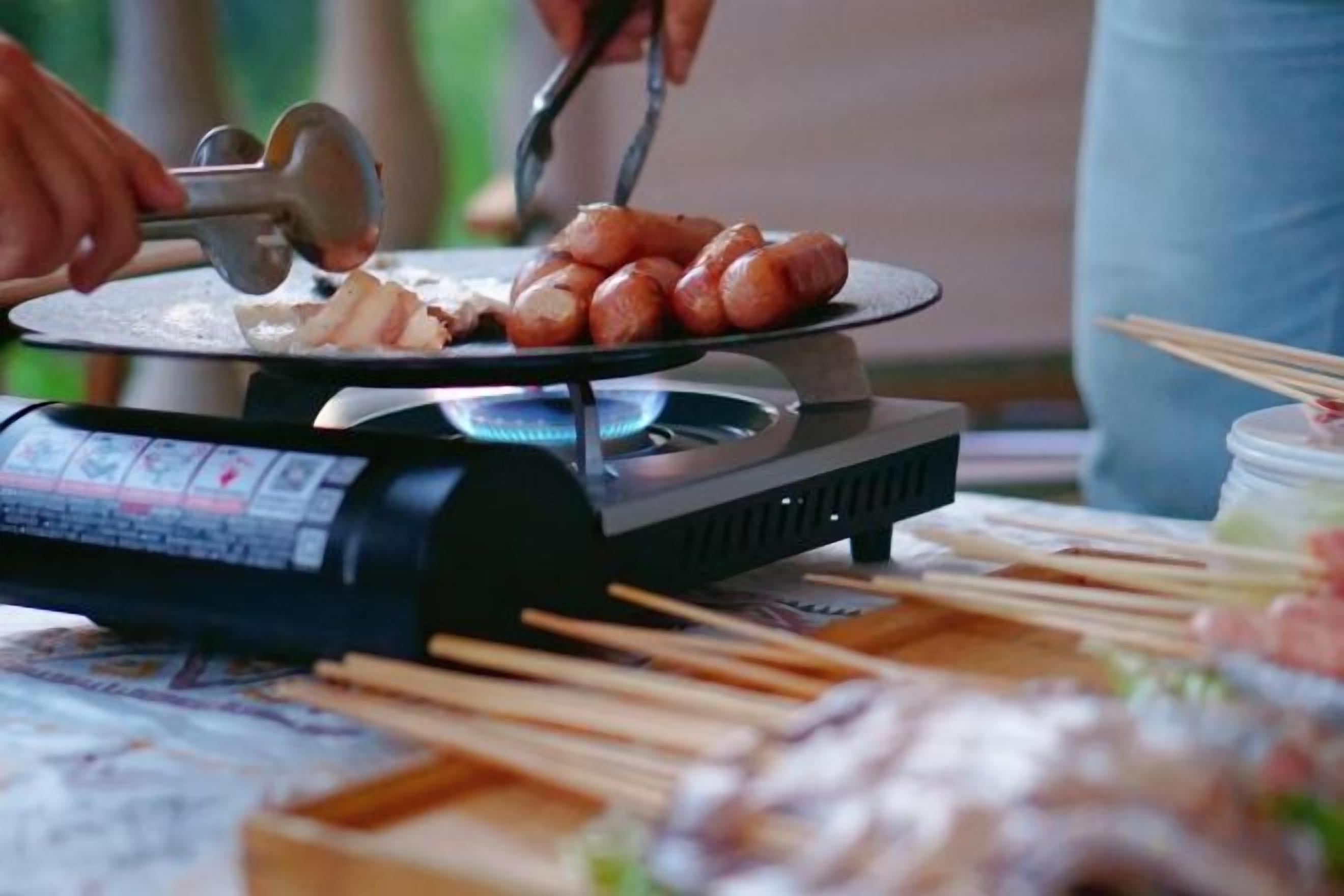 A portable grill with sausages being cooked, surrounded by skewers and a digital control panel.