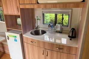 A compact kitchenette with a stainless steel sink, wooden cabinets, a window with a view of greenery, and a kettle on the counter. - To Journey Wuhu Longshan Campground (Wuhu)