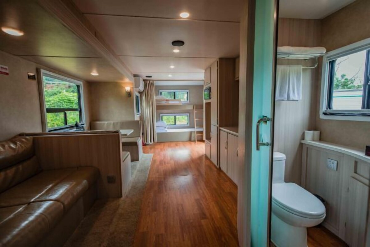 Un intérieur de camping-car compact avec un plancher en bois, un canapé en cuir marron, des toilettes blanches et une fenêtre donnant sur un paysage verdoyant.
