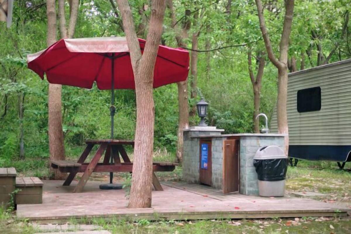 Een camping met een picknicktafel, een rode parasol, een mobiel toilet en een vuilnisbak.