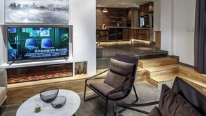 Una sala de estar moderna con televisor, asientos cómodos, una cocina al fondo y una escalera que sube a otro nivel.