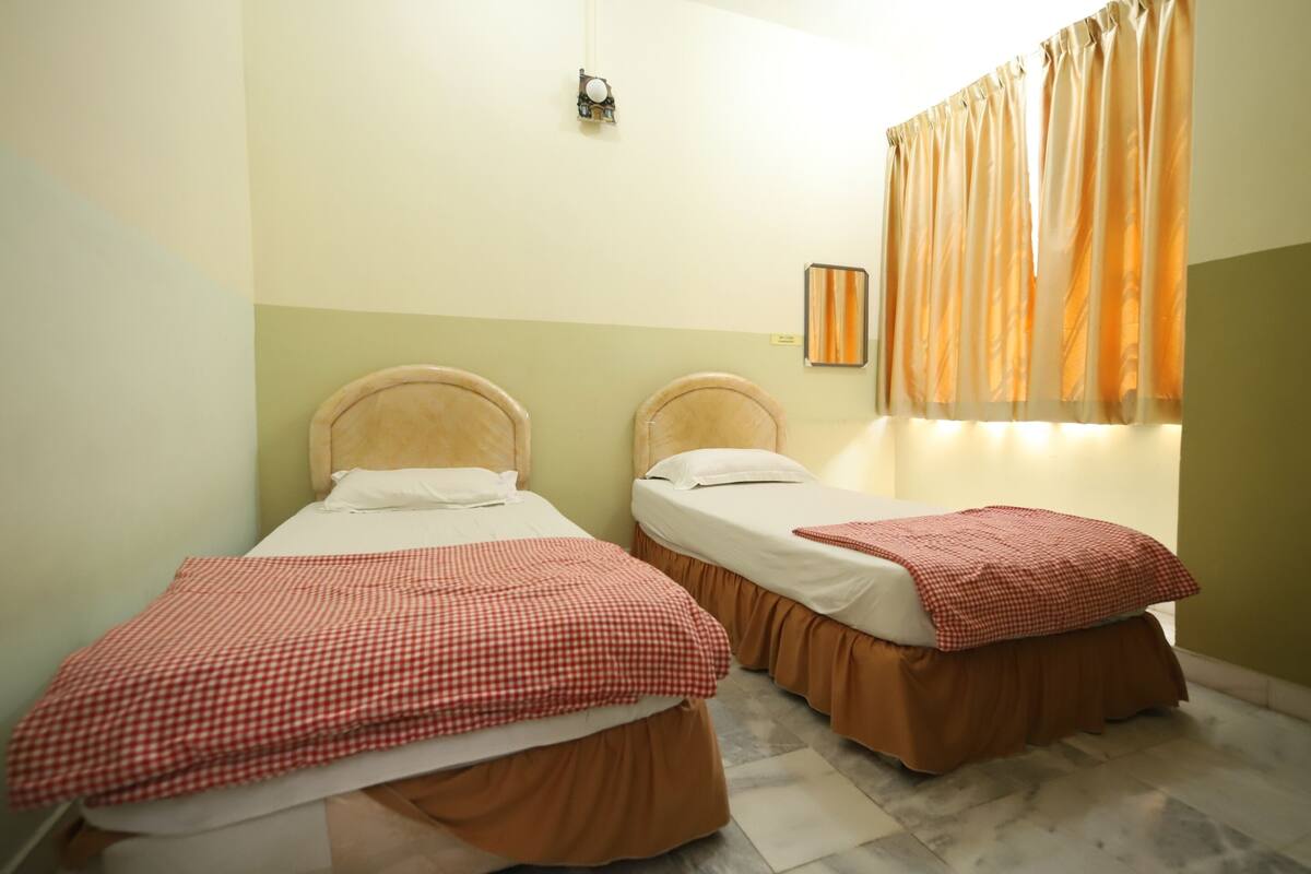 Una habitación con dos camas, cada una con una manta a cuadros y una falda marrón. Hay una ventana con cortinas naranjas y una lámpara fijada a la pared.