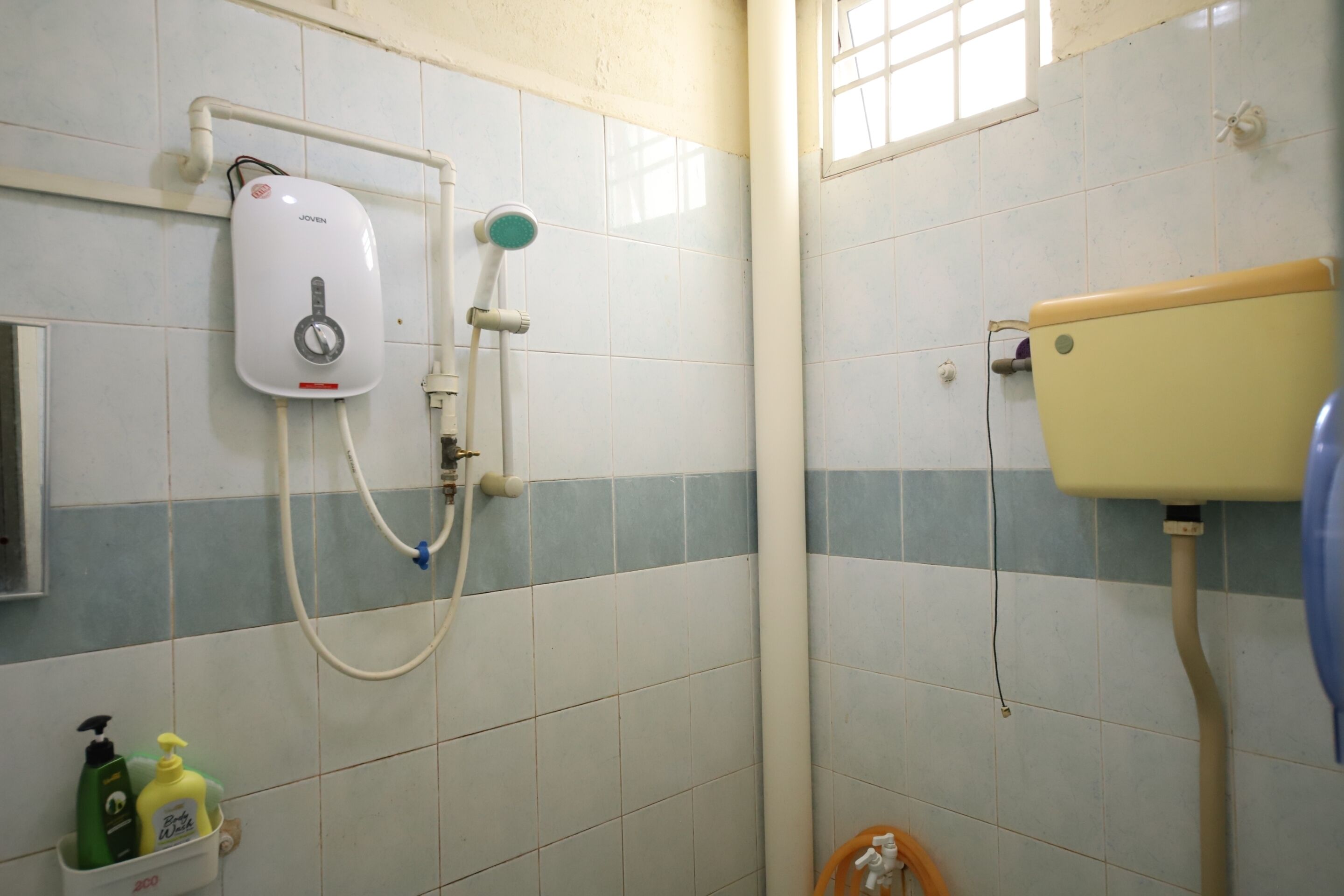 A bathroom with a white and blue tiled wall, a Joven water heater, a toilet, and a shower hose.