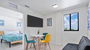 A modern hotel room with a bed, a dining area with chairs, a sofa, a TV, and a window with a city view.