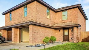Une maison à deux étages, construite en briques, avec un garage, une porte d’entrée et un petit jardin devant, composé d’une pelouse et de deux arbustes.