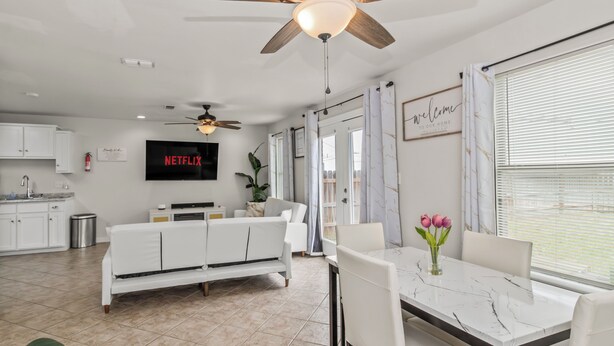 Un salon moderne avec un canapé blanc, une table à manger avec des chaises, un ventilateur de plafond et un téléviseur fixé au mur.